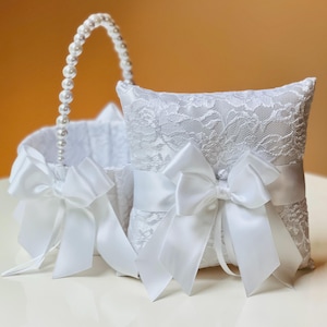 May include: A white lace ring pillow and a matching basket with a pearl handle. Both items are adorned with large white satin bows. The basket and pillow are designed for a wedding ceremony.