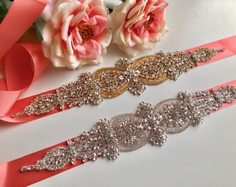 May include: Two bridal sashes with rhinestone embellishments. One sash is coral pink with a gold rhinestone design, the other is coral pink with a silver rhinestone design.