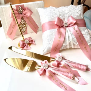 Może przedstawiać: Zestaw ślubny zawierający księgę gości, pióro, poduszkę na obrączki i zestaw do serwowania ciasta. Przedmioty zdobione są różowymi wstążkami, kryształowymi ozdobami i złotymi akcentami. Księga gości i wstążki mają niestandardowy tekst.