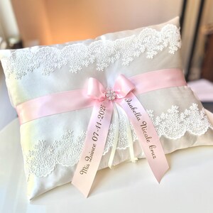 May include: White satin pillow with lace trim and pink satin ribbon. The ribbon is tied in a bow with a pearl embellishment. The ribbon is personalized with the text "Miss Quince 07-11-2025" and "Isabella Nicole Cruz".