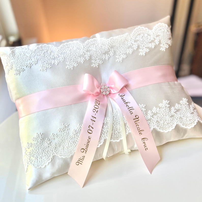 May include: White satin pillow with lace trim and pink satin ribbon. The ribbon is tied in a bow with a pearl embellishment. The ribbon is personalized with the text "Miss Quince 07-11-2025" and "Isabella Nicole Cruz".