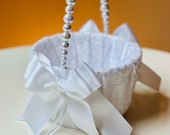 Op de afbeelding: Witte bloemenmeisjesmand met een parelhandvat en een grote satijnen strik. De mand is gevoerd met witte stof en heeft een delicate kanten overlay. Een bruiloft accessoire.