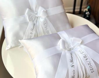 May include: Two white satin pillow ring bearer cushions with white satin ribbon and pearl trim. The ribbon is tied in a bow and has the names "Alexis & Sharon" and the date "05/24/2025" printed on it.
