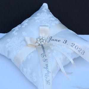 May include: A square white ring bearer cushion featuring a delicate lace overlay. A cream-coloured satin ribbon is tied in a bow at the centre, accented by a circular pearl and rhinestone brooch. The ribbon is printed with "K. Nycha & Damond" and "June 3, 2023."