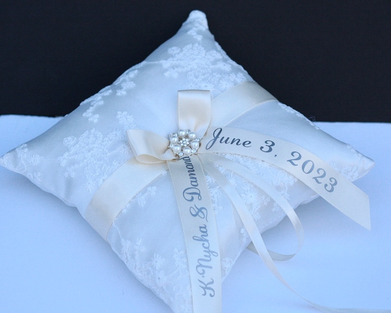May include: A square white ring bearer cushion featuring a delicate lace overlay. A cream-coloured satin ribbon is tied in a bow at the centre, accented by a circular pearl and rhinestone brooch. The ribbon is printed with "K. Nycha & Damond" and "June 3, 2023."