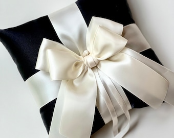May include: A black satin pillow with a large ivory satin bow. The pillow is designed for holding wedding rings.