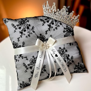 May include: A black lace ring pillow with a white ribbon and a silver tiara. The pillow has the text "Mis Quince Años" and "Lily 08/30/25" on the ribbon. The pillow is square and has a floral lace pattern.