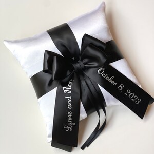 May include: White satin pillow with a black satin ribbon tied in a bow. The ribbon is personalized with the text "Lynne and Ron" and "October 8, 2023".