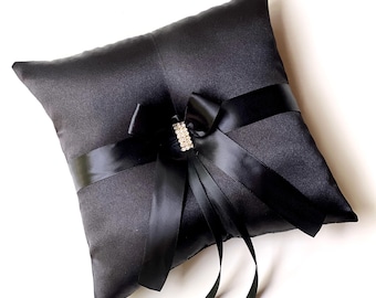 May include: A black satin ring pillow with a decorative black bow and a sparkling rhinestone accent. The pillow is square-shaped and features a satin ribbon that wraps around the center. A classic accessory for a wedding ceremony.