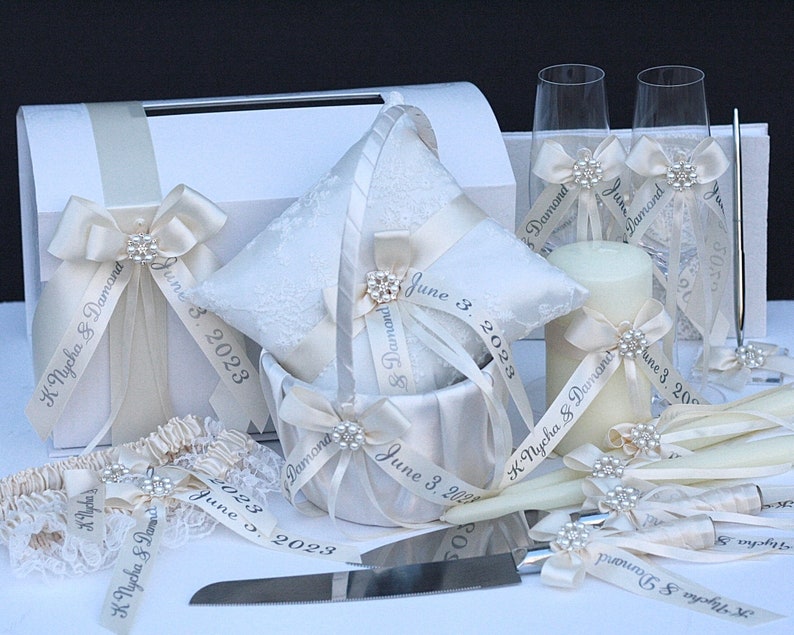 May include: A wedding accessory set featuring a white card box, a lace ring pillow, a flower basket, two champagne flutes, a pillar candle, a cake knife, and a garter. Each item is decorated with cream ribbons printed with K Nycha & Damond and June 3, 2023.