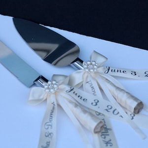 May include: A silver cake knife and server set with metallic blades and handles wrapped in cream-coloured ribbon. Each handle features a decorative pearl-encrusted flower accent and ribbons printed with the text June 3, 2023 and names.