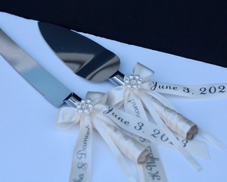 May include: A silver cake knife and server set with metallic blades and handles wrapped in cream-coloured ribbon. Each handle features a decorative pearl-encrusted flower accent and ribbons printed with the text June 3, 2023 and names.