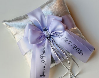 May include: A silver pillow with a lavender satin ribbon tied in a bow. The ribbon has the text "James & Patricia" and "01/27/2024" printed on it. The pillow is decorated with a silver beaded chain.