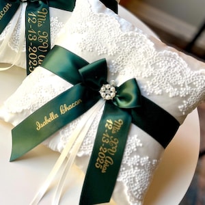Peut inclure: Deux coussins de porteur d'anneaux blancs avec des d&eacute;tails en dentelle. Chaque coussin est orn&eacute; d'un ruban vert fonc&eacute; avec du texte dor&eacute;, d'un n&oelig;ud d&eacute;coratif et d'une d&eacute;coration en strass. Le texte sur le ruban indique "Isabella Chacon" et "Mrs. & Mr. 12-13-2025".