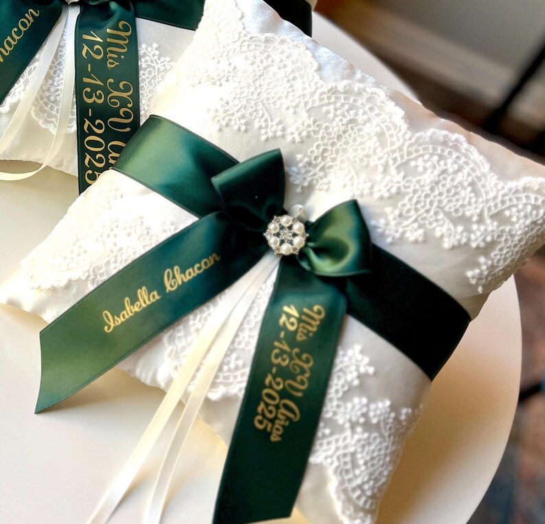 Peut inclure: Deux coussins de porteur d'anneaux blancs avec des d&eacute;tails en dentelle. Chaque coussin est orn&eacute; d'un ruban vert fonc&eacute; avec du texte dor&eacute;, d'un n&oelig;ud d&eacute;coratif et d'une d&eacute;coration en strass. Le texte sur le ruban indique "Isabella Chacon" et "Mrs. & Mr. 12-13-2025".