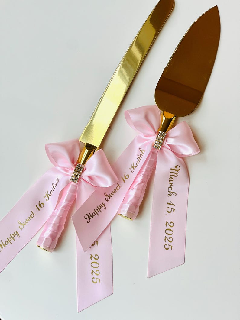 May include: Gold cake knife and server set with pink satin ribbons. The ribbons are tied in bows and have the text "Happy Sweet 16 Kailah" and "March 15, 2025" printed in gold.