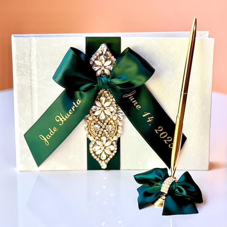 Peut inclure: Un livre d'or blanc avec un ruban vert et un stylo dor&eacute;. Le ruban porte les inscriptions "Jade Huerta" et "June 14, 2023". Le stylo est orn&eacute; d'un n&oelig;ud vert avec un accent en cristal.
