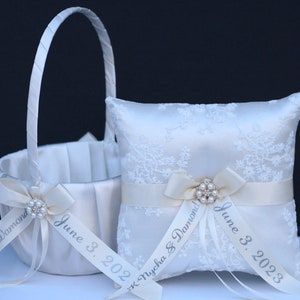 May include: A white satin flower girl basket and a matching square ring bearer pillow, both featuring white lace overlays, satin ribbons, and central pearl-encrusted brooches. The ribbons are printed with the text K. Nycha & Damond June 3, 2023.