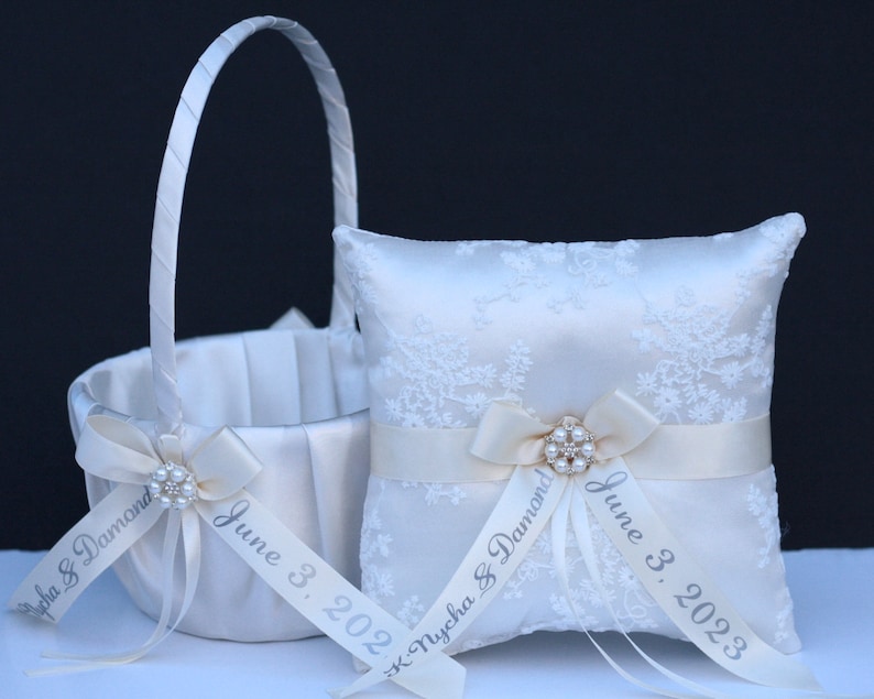 May include: A white satin flower girl basket and a matching square ring bearer pillow, both featuring white lace overlays, satin ribbons, and central pearl-encrusted brooches. The ribbons are printed with the text K. Nycha & Damond June 3, 2023.