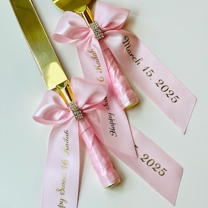 May include: Two gold cake cutting knives with pink satin ribbon bows and gold lettering. The ribbon on one knife says "Happy Sweet 16 Kailah" and the other says "March 15, 2025 16 Kailah".