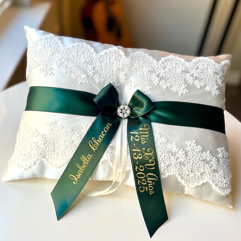 Peut inclure: Un coussin d'alliances en satin blanc avec une superposition de dentelle. Un ruban vert fonc&eacute; est nou&eacute; autour du coussin avec un n&oelig;ud et un bijou &eacute;tincelant. Deux rubans verts avec du texte dor&eacute; sont attach&eacute;s. L'un dit "Isabella Chacon" et l'autre "Mis 15 A&ntilde;os 12-13-2025".