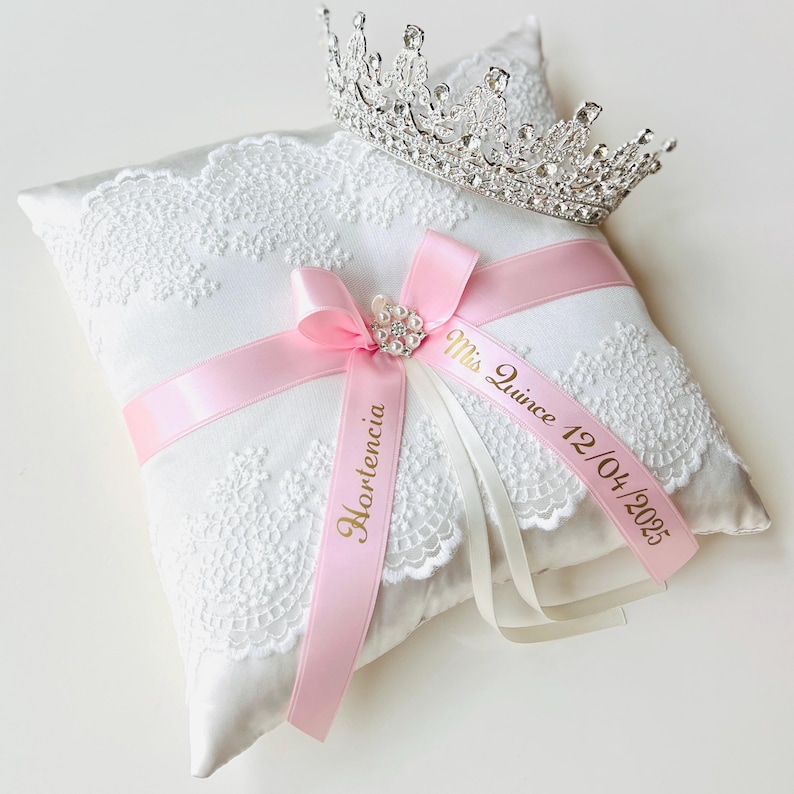 May include: A white satin pillow with lace trim and pink satin ribbon tied in a bow. The ribbon has the text "Miss Quince 12/04/2025" and "Hortensia" printed in gold. A silver tiara with rhinestones sits on top of the pillow.