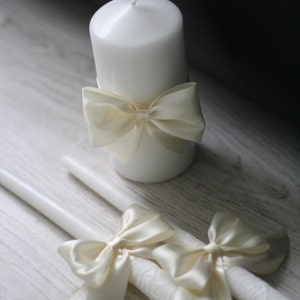 May include: Two white candles with ivory satin bows. The candles are on a white wooden surface.