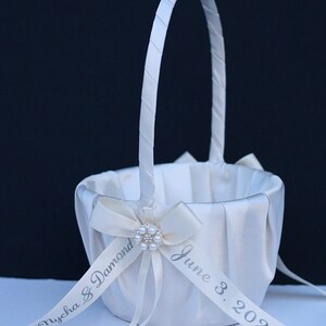 May include: A white satin flower girl basket with a tall, wrapped handle. The basket features a pleated fabric exterior and a central cream-coloured ribbon bow adorned with a faux pearl brooch. A ribbon is printed with Nycha & Damond June 3, 2023.