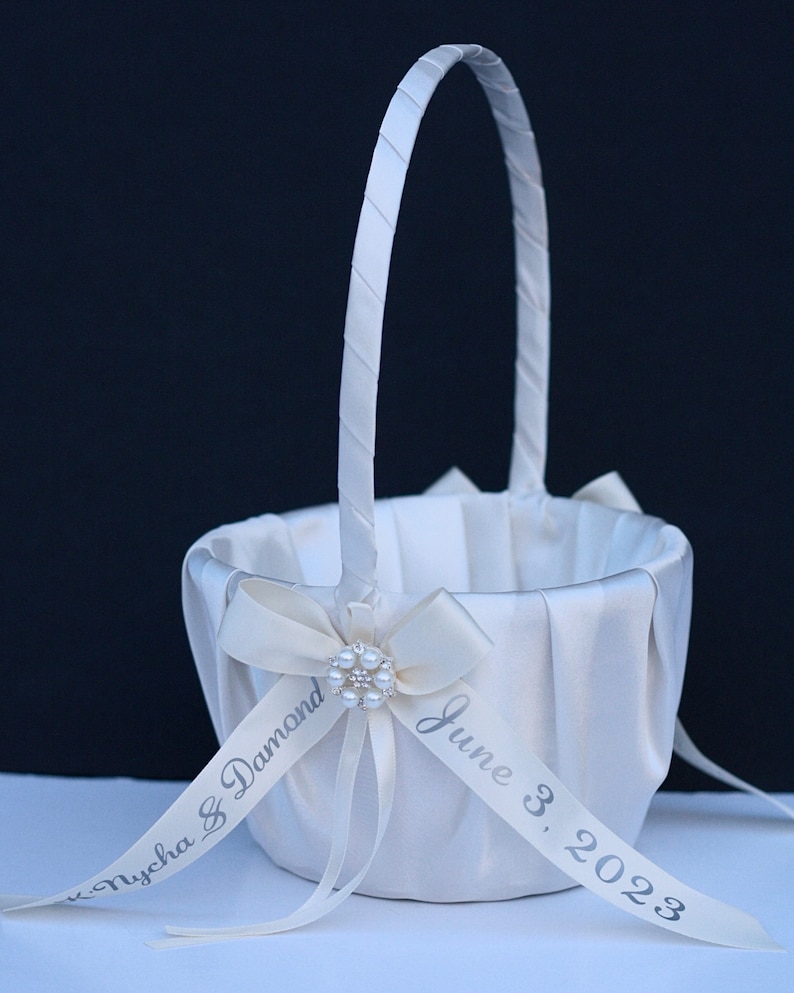 May include: A white satin flower girl basket with a tall, wrapped handle. The basket features a pleated fabric exterior and a central cream-coloured ribbon bow adorned with a faux pearl brooch. A ribbon is printed with Nycha & Damond June 3, 2023.