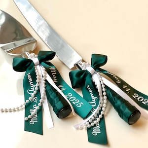 Peut inclure: Un ensemble de couteau et de pelle &agrave; g&acirc;teau argent&eacute;s. Les manches sont envelopp&eacute;s d'un ruban de satin vert fonc&eacute; avec le texte blanc "May 11, 2025" et "Marka & Aymen". Des guirlandes de perles blanches et des n&oelig;uds ornent les manches.