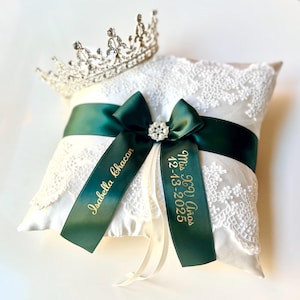 Peut inclure: Un coussin d'alliances en satin blanc avec une superposition de dentelle, orn&eacute; d'un ruban et d'un n&oelig;ud vert fonc&eacute;. Le n&oelig;ud est orn&eacute; d'une broche en strass. Le coussin porte l'inscription dor&eacute;e "Isabella Chacon" et "Mis XV A&ntilde;os 12-13-2025". Une tiare argent&eacute;e est pos&eacute;e dessus.