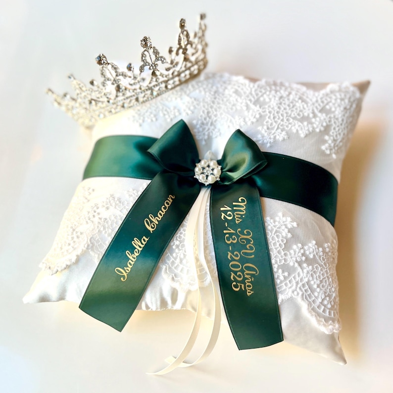 Peut inclure: Un coussin d'alliances en satin blanc avec une superposition de dentelle, orn&eacute; d'un ruban et d'un n&oelig;ud vert fonc&eacute;. Le n&oelig;ud est orn&eacute; d'une broche en strass. Le coussin porte l'inscription dor&eacute;e "Isabella Chacon" et "Mis XV A&ntilde;os 12-13-2025". Une tiare argent&eacute;e est pos&eacute;e dessus.