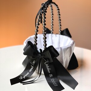 May include: White satin flower girl basket with a pearl handle and black satin ribbon bows. The ribbon is personalized with the text "Lynne and Jon" and "October 8, 2023".