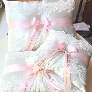 May include: Two white satin pillow cushions with lace trim and pink satin ribbon tied with a bow. The ribbon has gold lettering that reads "Mis Quince 07-11-2023" and "Isabella Nicole Cruz".