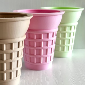 May include: Three colorful ice cream cone-shaped planters. The planters are in shades of beige, pink, and green. The cone portion has a waffle pattern, and the top is a smooth, rounded bowl. The planters are arranged in a row.