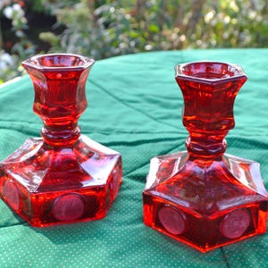 May include: Two red glass candle holders. Each has a hexagonal base with circular accents and a flared top. The candle holders are made of a transparent red glass and sit on a green surface.