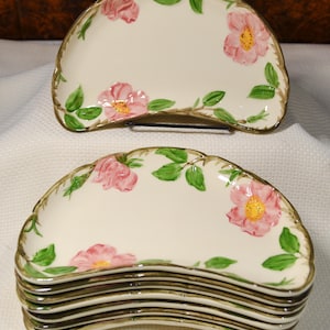 May include: A set of six crescent-shaped ceramic plates featuring a cream-colored base with hand-painted pink wild roses, green leaves, and brown stems. Each plate has a scalloped edge with a gold-toned trim along the curved outer rim.