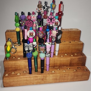 May include: A tiered wooden stand with multiple holes to hold a variety of colorful pens with decorative tops. The tops feature various designs, including animals, words, and patterns. Some of the words on the tops include "Know Worth", "Easily Distracted", "Just a Little Moody", and "Ask Me About My Lashes".
