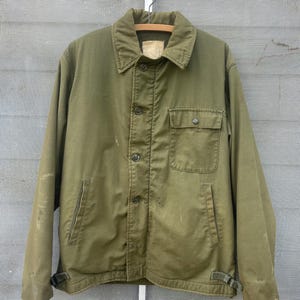 May include: An olive green jacket with a button-up front, a collar, and two front pockets. The jacket has a worn appearance, with some discoloration and wear visible. It is hanging on a metal hanger.