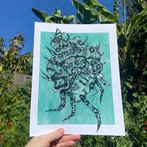 May include: A teal and black print of a woman with a head full of eyes and tentacles. The woman's face is serene and she is looking down. The print is on a light teal background.