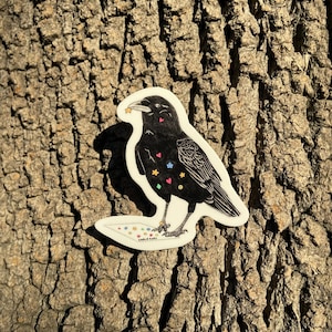 Puede incluir: Una pegatina de cuervo negro con corazones, estrellas y otras formas coloridas en su cuerpo. El cuervo sostiene una estrella en su pico y está de pie sobre un papel blanco con el texto "Charlie Rains" en él.