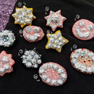 Könnte beinhalten: Zehn bunte Harz-Pins in verschiedenen Formen und Designs. Die Pins haben eine weiße, silberne und schimmernde Glitzer-Oberfläche. Die Pins sind in Form von Sternen, Smiley-Gesichtern und anderen abstrakten Formen.