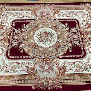 May include: A large, rectangular area rug with a detailed floral design. The rug features a deep red background with cream, beige, and gray accents. The central design is a floral medallion, surrounded by ornate borders and floral patterns.
