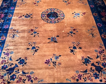 Op de afbeelding: Een vintage Chinees tapijt met een donkerblauwe rand en een centraal medaillon. Het tapijt heeft een bloemenpatroon met vogels en een lichtbruine achtergrond.