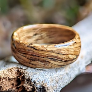 Peut inclure: Une bague en bois avec un motif de grain de bois naturel et une finition brillante. La bague est brune avec des stries brun foncé et repose sur un morceau de bois flotté.