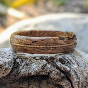 Peut inclure: Une bague en bois faite à la main avec un design unique. La bague présente une bande de bois brun clair avec des marbrures marron foncé et noires. Un fin fil métallique enroulé est intégré au centre de la bague. La bague est présentée sur un morceau de bois flotté.