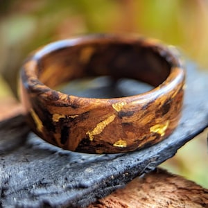 Peut inclure: Une bague en bois avec des paillettes dorées incrustées dans le bois. La bague est brune et noire avec un motif de grain de bois naturel.
