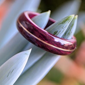Peut inclure: Gros plan d'une bague en bois de couleur pourpre foncé et or avec une finition polie. La bague est posée sur une plante verte et blanche.