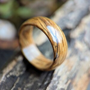 May include: A wooden ring with a natural wood grain pattern and a clear epoxy finish. The ring is brown with dark brown streaks and a light brown center.
