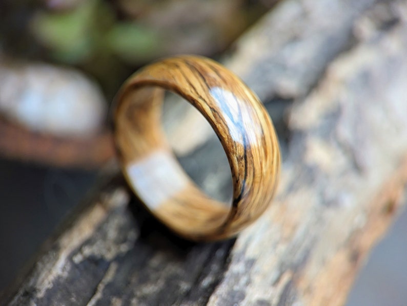 May include: A wooden ring with a natural wood grain pattern and a clear epoxy finish. The ring is brown with dark brown streaks and a light brown center.